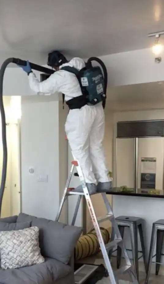 Technician on a ladder using a backpack vacuum on the ceiling of an apartment
